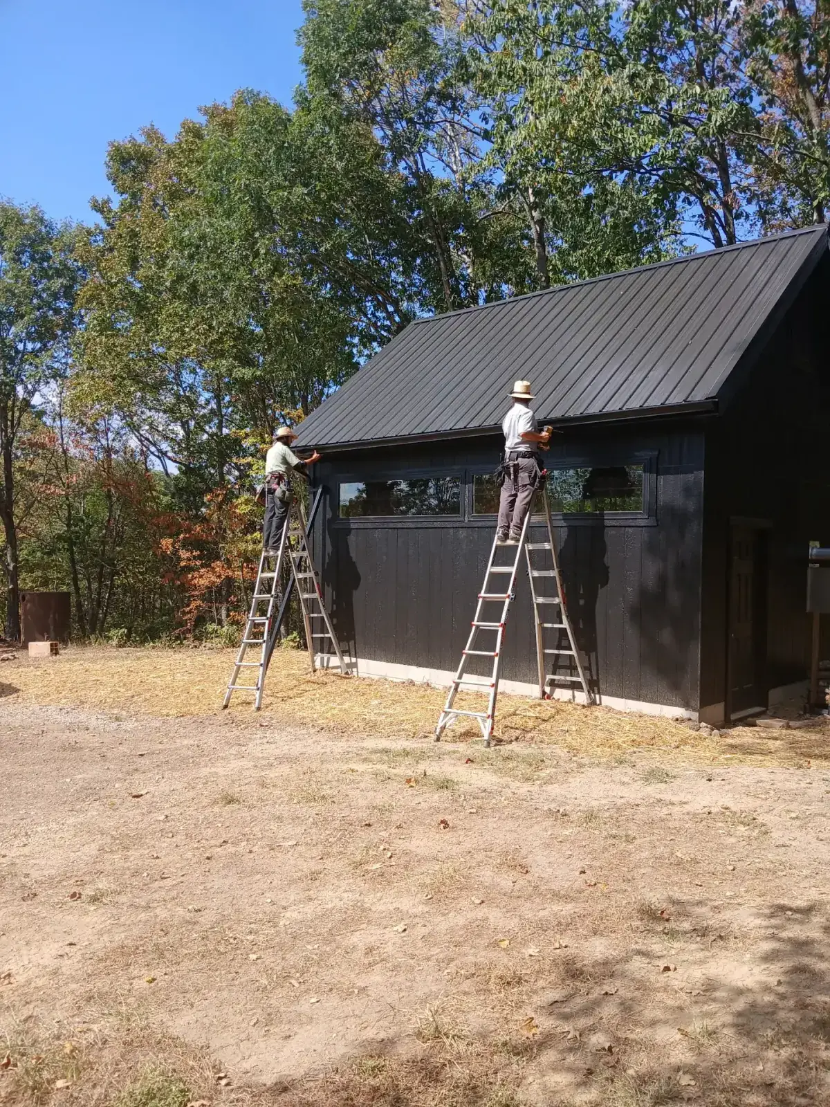 Two Amish roofers on ladders fitting trim to the gable of a black metal-roofed outbuilding